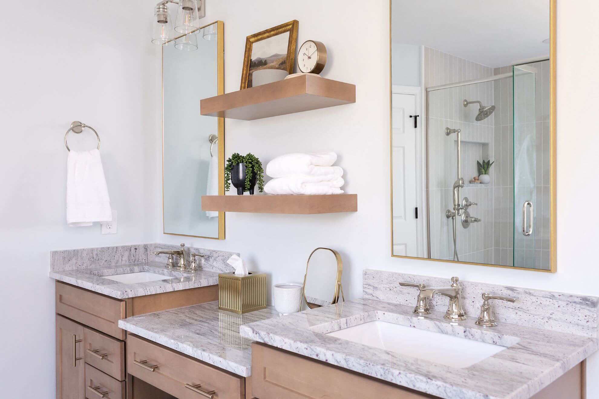 Master bathroom double vanity with granite countertops, gold-framed mirrors, natural wood floating shelves, and glass shower enclosure