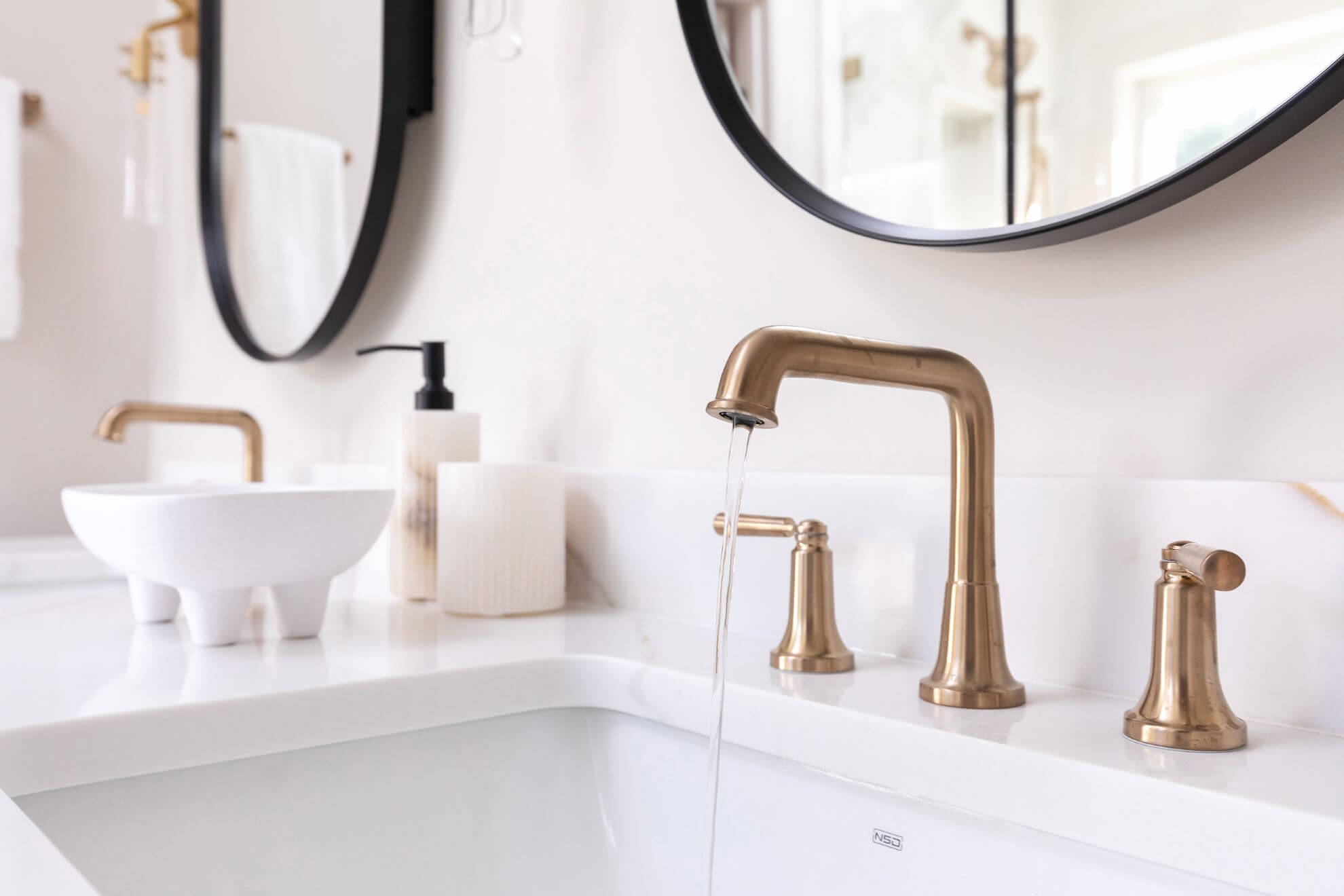 Close-up of Morrisville bathroom vanity featuring champagne bronze widespread faucet, white quartz countertop, and matte black oval mirrors