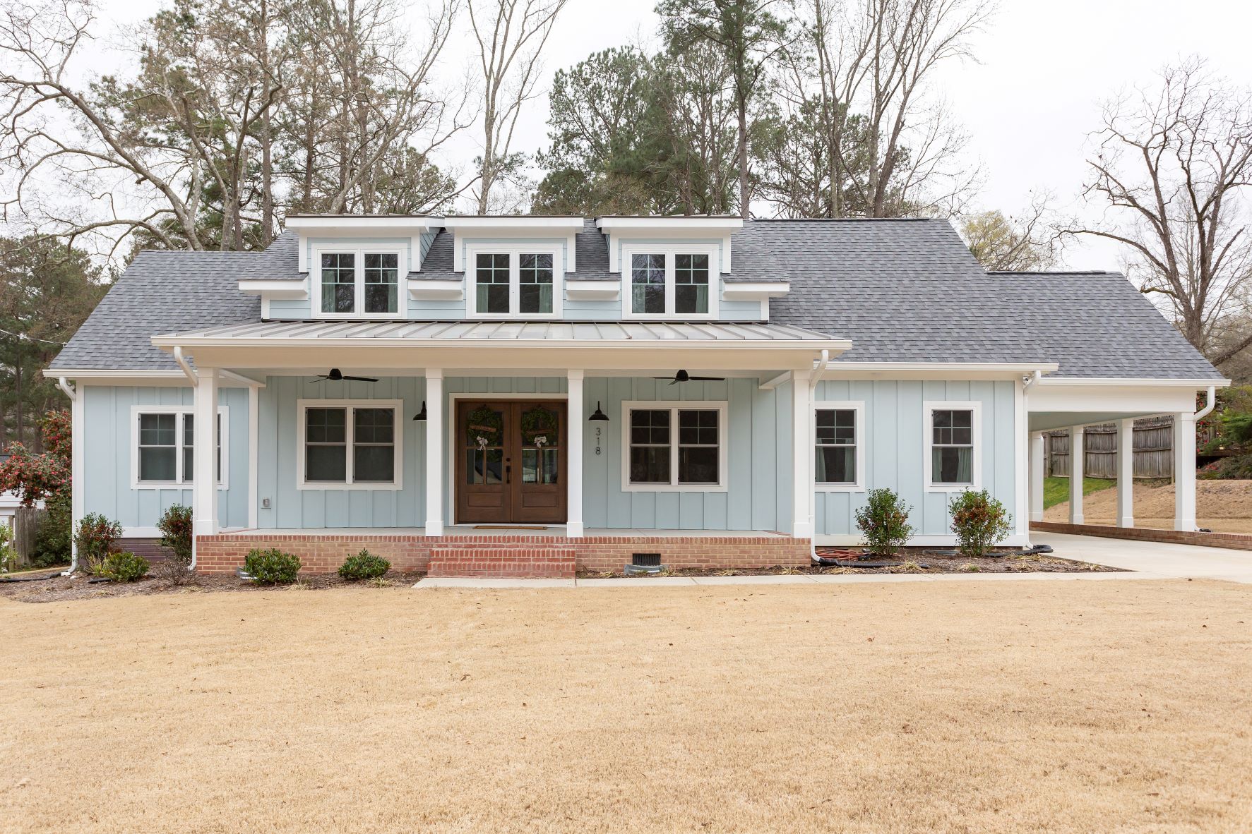 Custom farmhouse new construction with light blue board-and-batten siding, covered front porch, dormer windows, metal roof accent, wood double entry doors, and attached carport