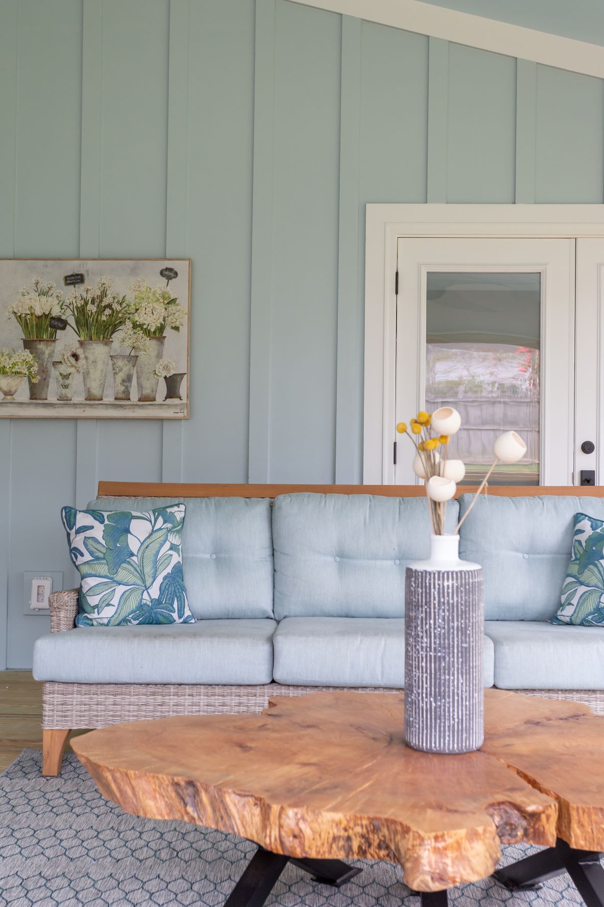 Fuquay-Varina screened patio with sage green board-and-batten walls, wicker outdoor sofa, live-edge wood coffee table, tropical pillows, and botanical artwork