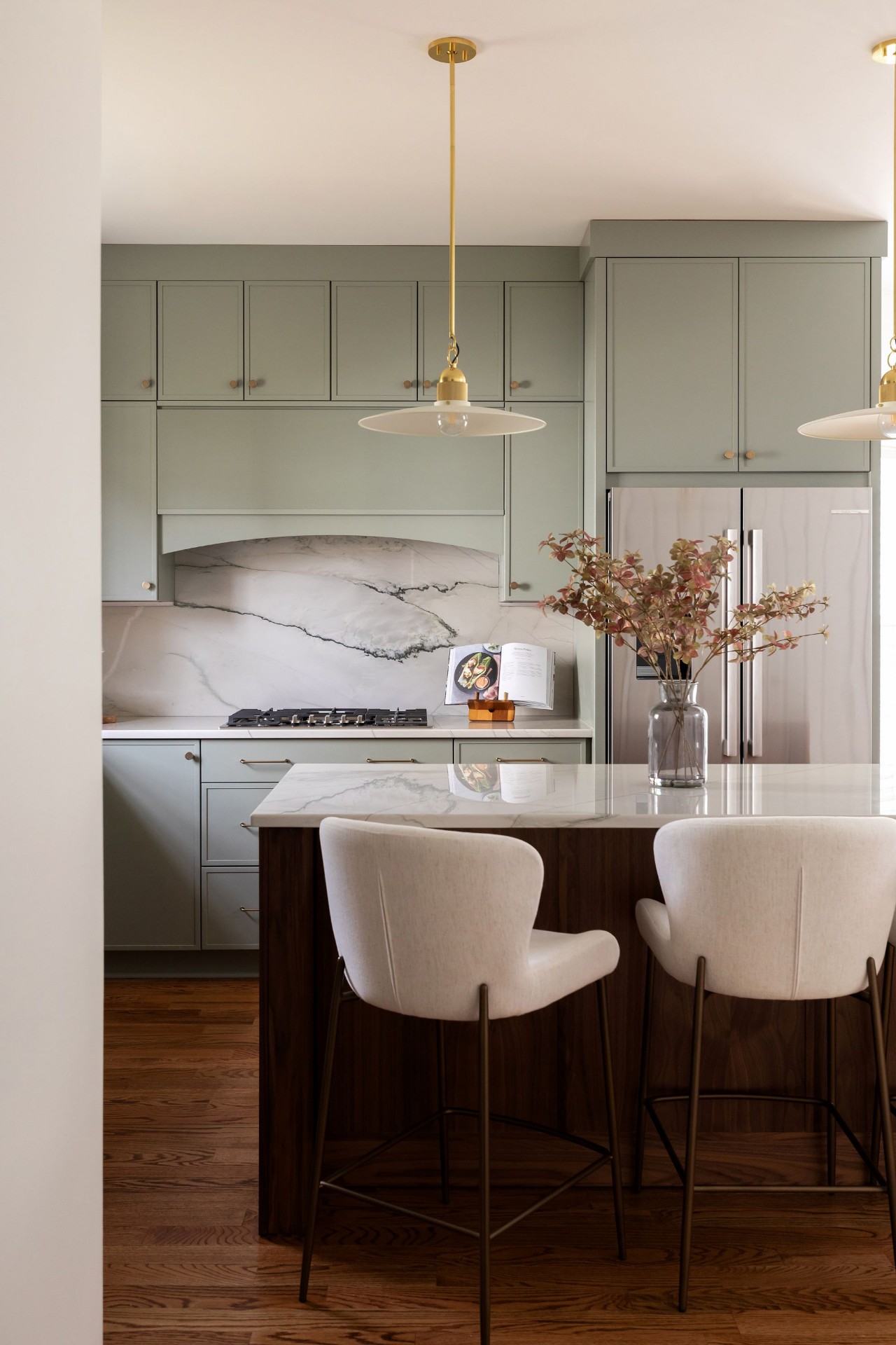 Designer Cary kitchen with veined marble slab backsplash, gas cooktop, sage green cabinets, and modern upholstered bar stools