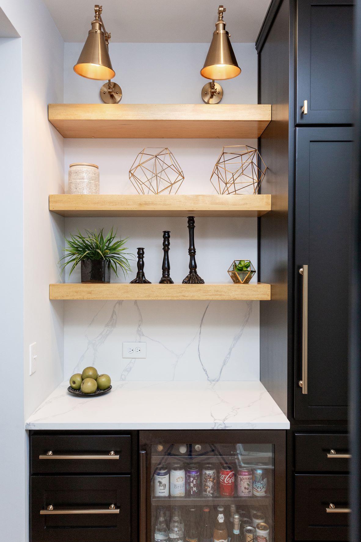Elegant butler's pantry with natural wood floating shelves, brass wall sconces, marble backsplash, black cabinets, and beverage refrigerator