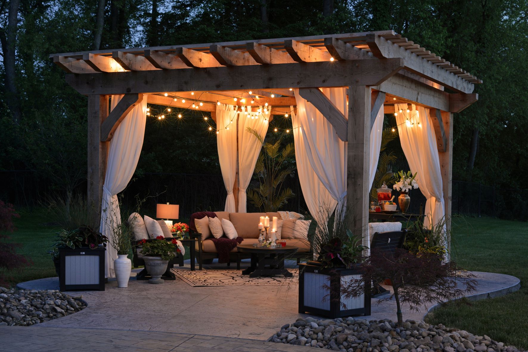 Evening outdoor living space featuring cedar pergola with string lights, sheer curtains, outdoor sofa, stamped concrete patio, and landscaping